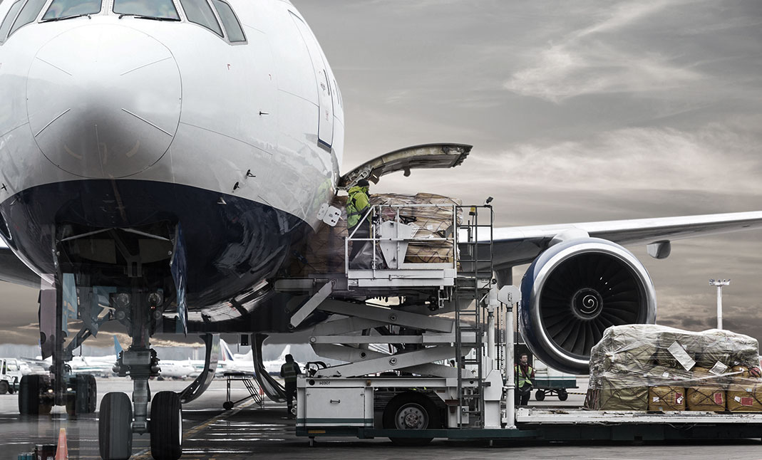 Ground Support Equipment (GSE) supporting the cargo goods loading operations of a container and a pallet of an aircraft on the ground of the airport tarmac thanks to a Container Loader. Plane lateral door is opened to allow the goods to get in. A men brings the load of goods to be accomodated inside the aircraft trunk